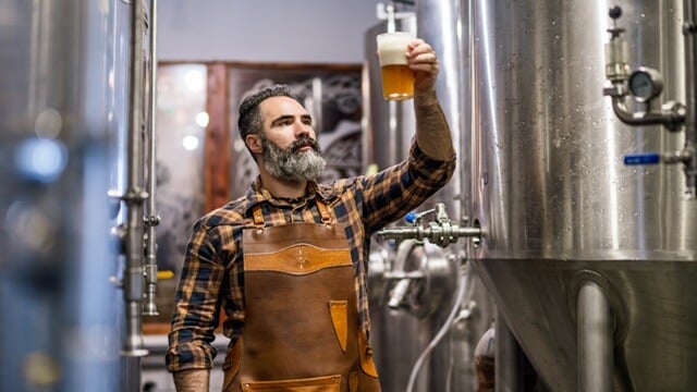 Smiling brewer holding a glass of beer in a brewery, surrounded by brewing equipment.